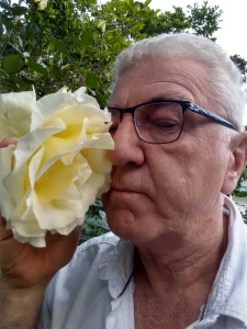 A close-up, vertical photograph of an older man with short white hair and black-rimmed glasses, gently holding and leaning in to smell a large, blooming pale yellow rose. Green foliage from a garden fills the background.